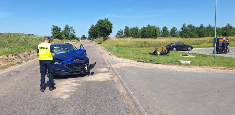 Miejsce wypadku drogowego wielu na Kaszubach. Na drodze stoi niebieski samochód z rozbitym przodem, obok którego stoi policjant. Na trawiastym poboczu po prawej stronie leży przewrócony motocykl. W tle widać zieleń i zaparkowane auto