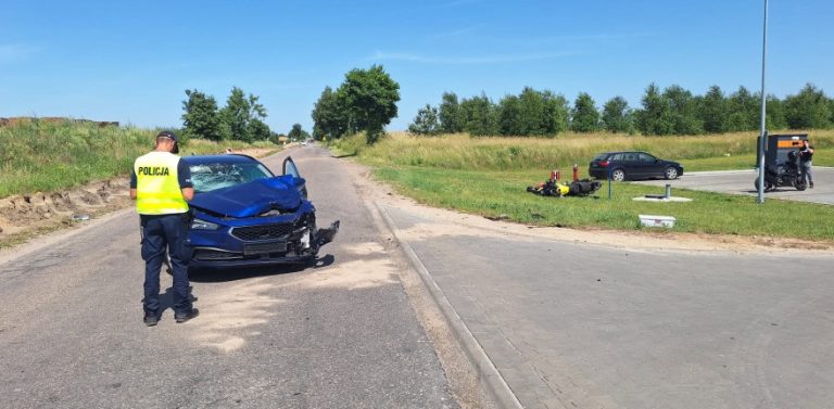 Miejsce wypadku drogowego wielu na Kaszubach. Na drodze stoi niebieski samochód z rozbitym przodem, obok którego stoi policjant. Na trawiastym poboczu po prawej stronie leży przewrócony motocykl. W tle widać zieleń i zaparkowane auto