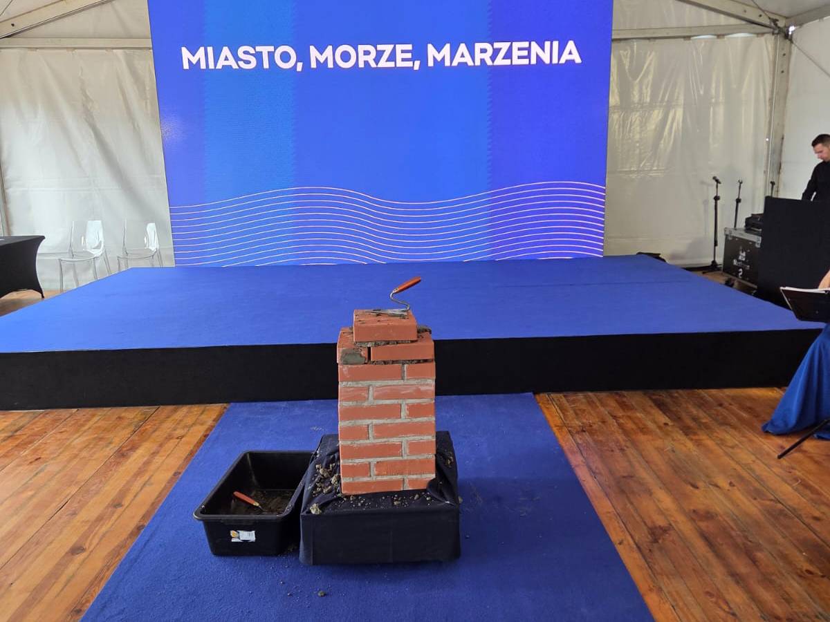 Wmurowanie kamienia węgielnego pod budowę kompleksu Nova Marina w Gdyni. Na pierwszym planie, centralnie, znajduje się stos cegieł ułożonych w kształt niewielkiego murku, na którym leży kielnia budowlana z drewnianą rączką. Cegły stoją na czarnej podstawce, obok nich widać czarną kuwetę z zaprawą murarską.Cała ta konstrukcja znajduje się na niebieskim podwyższeniu, prawdopodobnie podium lub platformie. Podłoga wokół to drewniane deski.W tle widoczny jest baner z napisem wielkimi literami: "MIASTO, MORZE, MARZENIA", umieszczonym na tle błękitnego gradientu. Po prawej stronie ekranu widać sylwetkę mężczyzny w ciemnym garniturze, a po lewej puste, przezroczyste krzesła. Całość sugeruje uroczysty charakter wydarzenia związanego z rozpoczęciem nowej inwestycji