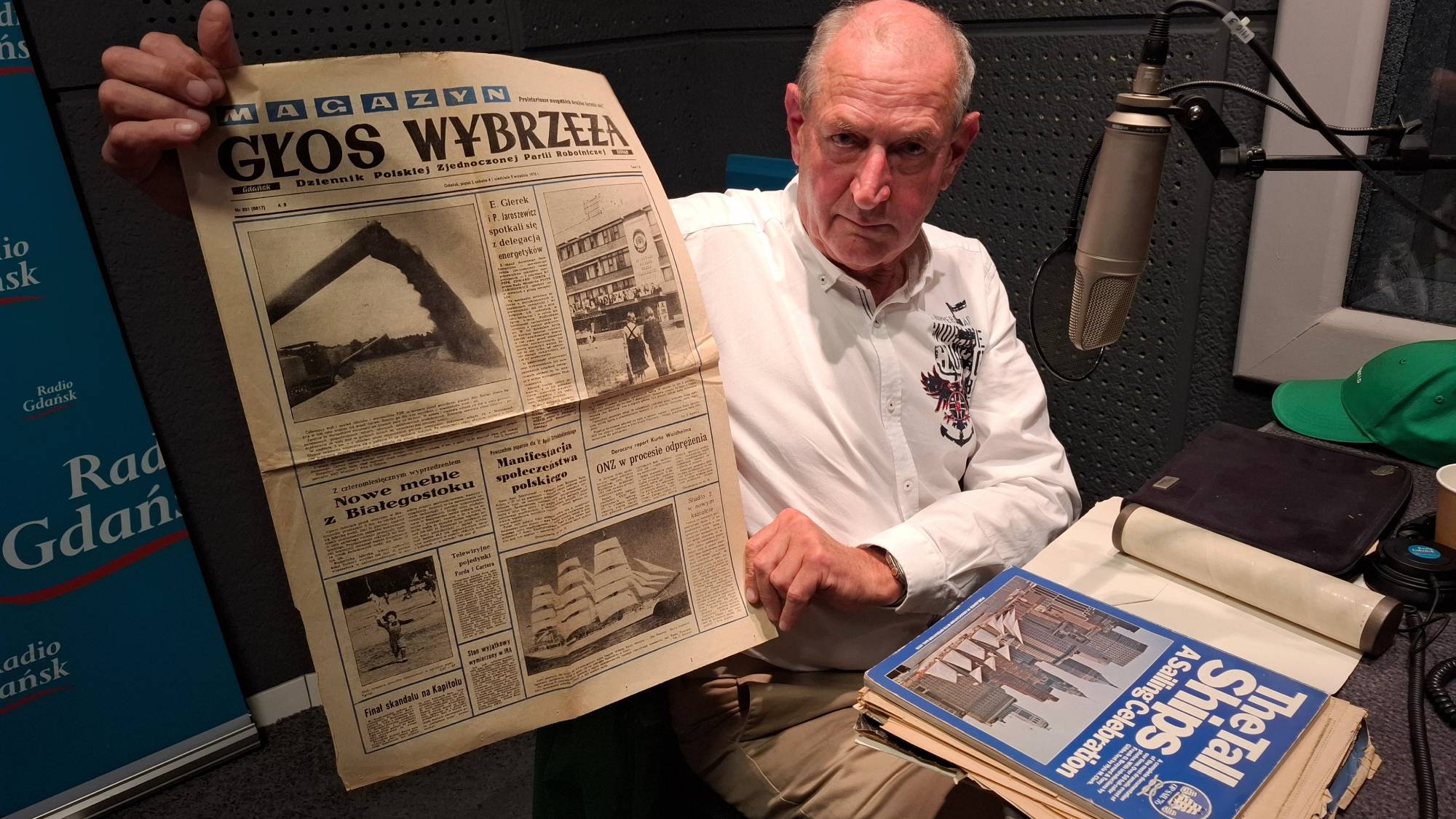 Kapitan Maciej Polarczyk w studiu radiowym trzyma archiwalne wydanie gazety „Głos Wybrzeża”, prezentując je do aparatu. W gazecie jest zdjęcie Daru Pomorza ilustrujące artykuł o rejsie na 200-lecie Stanów Zjednoczonych. Na biurku obok mikrofonu leży album o żaglowcach „The Tall Ships”