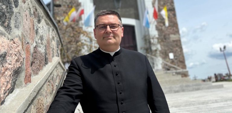 Ksiądz Przemysław Szulc stojący na zewnątrz kościoła z kamienną ścianą i schodami w tle