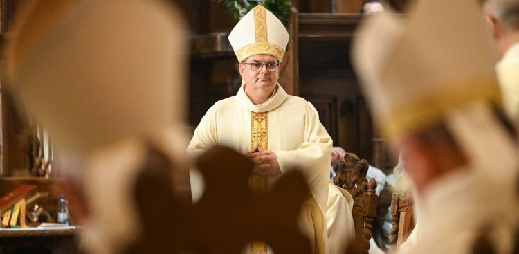 Ks. Przemysław Szulc w jasnym ornacie i mitrze na głowie, ze złożonymi dłońmi, podczas uroczystości święceń biskupich w katedrze w Pelplinie. Zdjęcie wykonane zza ramion innych duchownych, których postacie na pierwszym planie są rozmyte