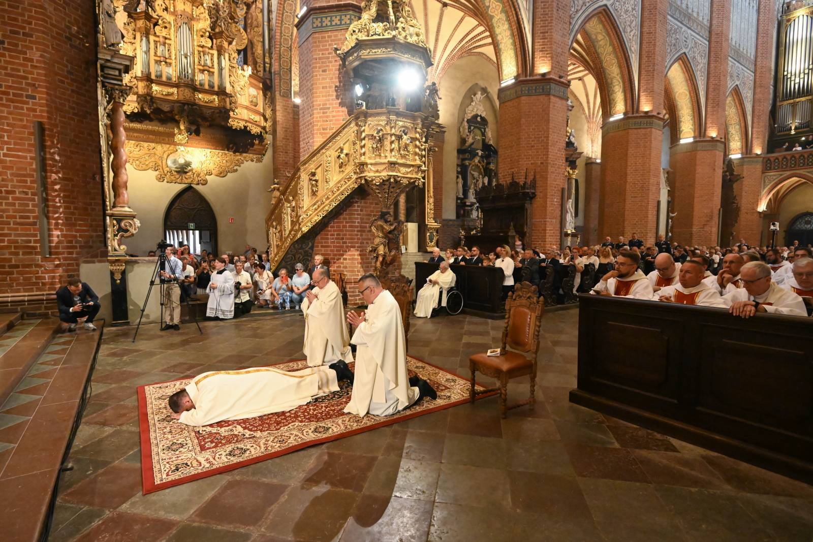 Moment prostracji (leżenia krzyżem) ks. Przemysława Szulca na dywanie przed ołtarzem podczas święceń biskupich w Bazylice Katedralnej w Pelplinie. Obok niego klęczą dwaj księża asystujący, a w tle widać monumentalne, ceglane wnętrze świątyni, zdobioną ambonę oraz zgromadzonych uczestników liturgii