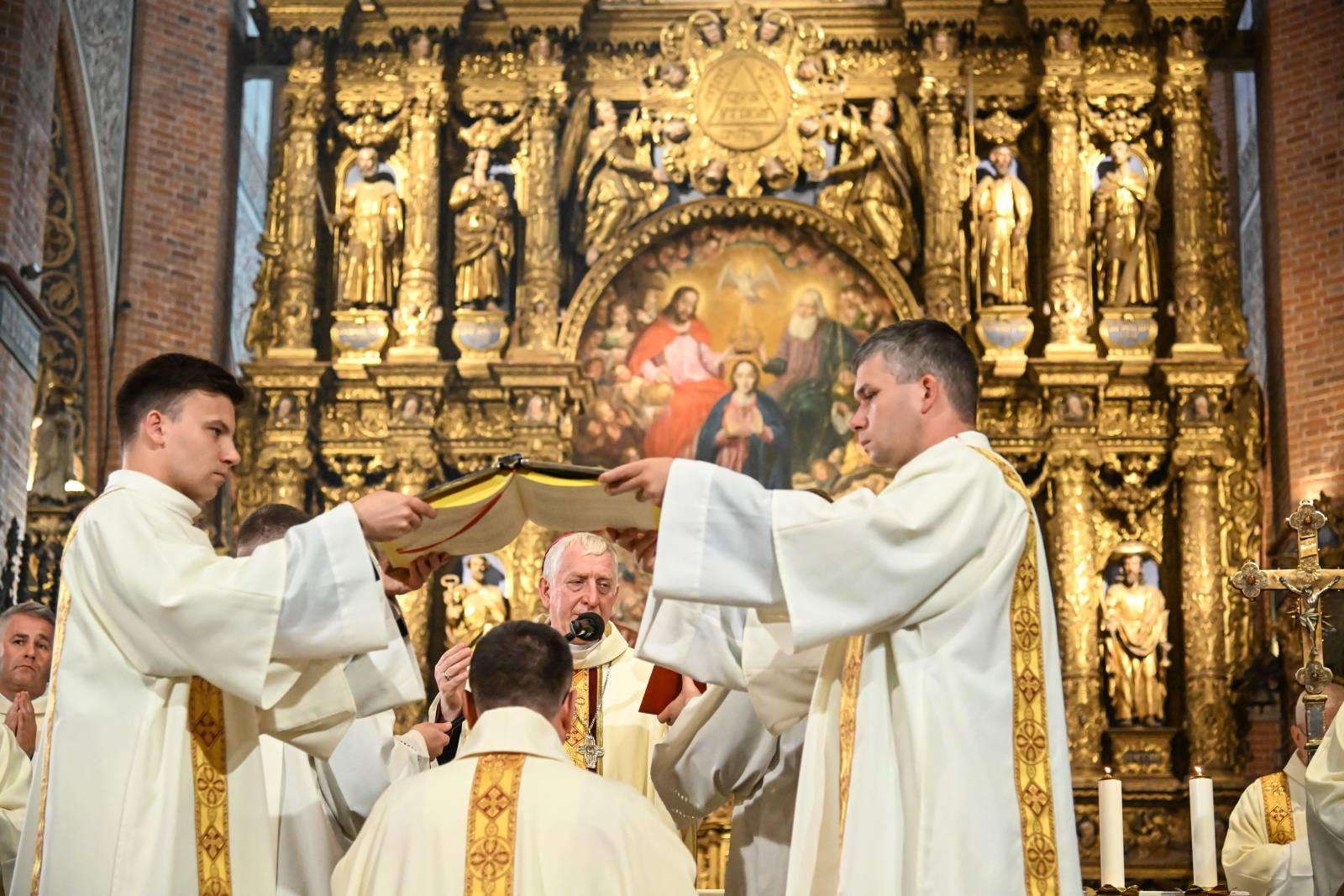 Obrzęd nałożenia otwartej księgi Ewangelii (Ewangelarza) nad głową klęczącego ks. Przemysława Szulca podczas jego święceń biskupich. Księgę trzyma dwóch duchownych w jasnych szatach liturgicznych. W tle widoczny jest celebrujący biskup oraz monumentalny, złocony ołtarz główny katedry w Pelplinie