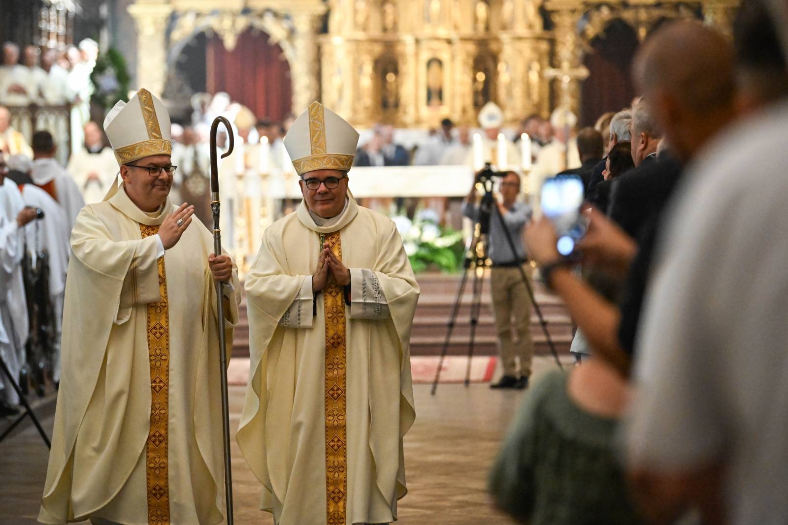 Nowo wyświęcony bp Przemysław Szulc oraz biskup pelpliński Ryszard Kasyna idą nawą główną katedry w Pelplinie podczas procesji. Bp Szulc uśmiecha się, trzyma pastorał i pozdrawia wiernych gestem dłoni, a bp Kasyna kroczy obok niego z dłońmi złożonymi do modlitwy. W tle widać zgromadzonych ludzi, z których część robi zdjęcia telefonami