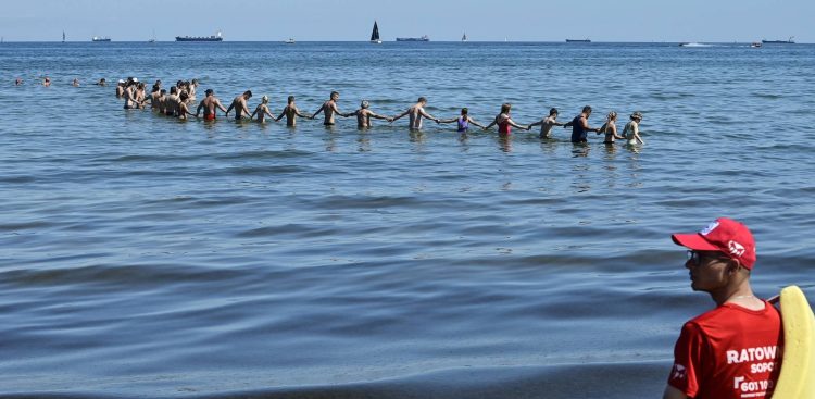Łańcuch życia utworzony w morzu przez plażowiczów i ratowników. W prawym dolnym rogu, na pierwszym planie, widoczny jest z profilu ratownik wodny w czerwonej koszulce z napisem „RATOWNIK” oraz w czerwonej czapce z daszkiem, trzymający żółtą boję ratunkową. W głębi, w błękitnej wodzie sięgającej do pasa, kilkadziesiąt osób w strojach kąpielowych stoi w długiej linii i trzyma się za ręce, tworząc tyralierę przeszukującą dno morza. Na horyzoncie, pod bezchmurnym niebem, widać spokojną taflę wody z kilkoma statkami handlowymi i żaglówkami.