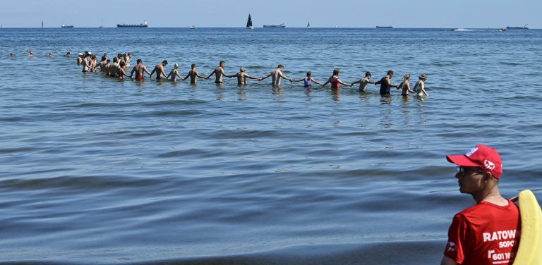 Łańcuch życia utworzony w morzu przez plażowiczów i ratowników. W prawym dolnym rogu, na pierwszym planie, widoczny jest z profilu ratownik wodny w czerwonej koszulce z napisem „RATOWNIK” oraz w czerwonej czapce z daszkiem, trzymający żółtą boję ratunkową. W głębi, w błękitnej wodzie sięgającej do pasa, kilkadziesiąt osób w strojach kąpielowych stoi w długiej linii i trzyma się za ręce, tworząc tyralierę przeszukującą dno morza. Na horyzoncie, pod bezchmurnym niebem, widać spokojną taflę wody z kilkoma statkami handlowymi i żaglówkami.