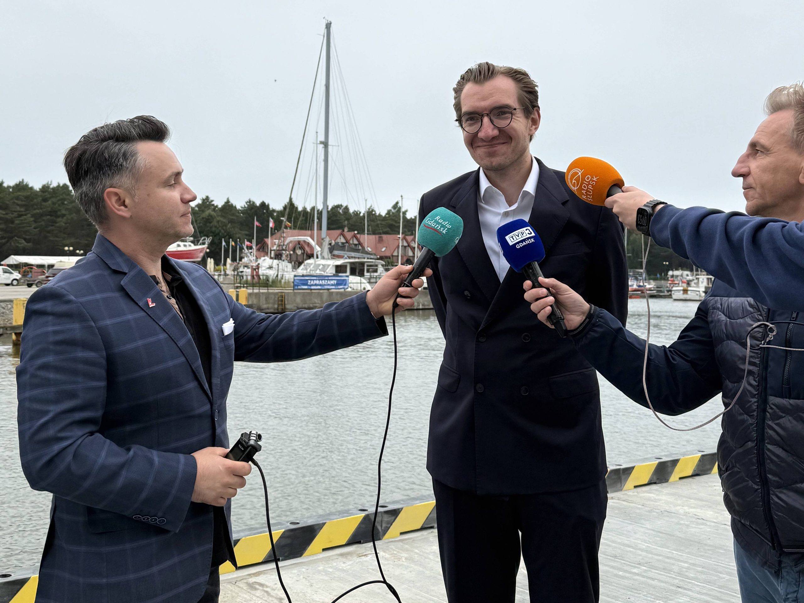 Zdjęcie przedstawia Jana Szyszko, Wiceministra Funduszy i Polityki Regionalnej, w trakcie konferencji prasowej w łebskim porcie, otoczonego przez dziennikarzy. Minister stoi w centrum, ubrany w ciemny garnitur i białą koszulę, uśmiecha się lekko i patrzy w prawo.Z obu stron podchodzą do niego dziennikarze, trzymając w rękach mikrofony z logotypami różnych mediów. Po lewej stronie, w granatowej marynarce z mikrofonem "Radio Gdańsk" skierowanym w stronę ministra reporter Łukasz Kosik. Po prawej stronie, mężczyzna w ciemnej kurtce, trzyma dwa mikrofony – jeden z logo "TVP3 Gdańsk" i drugi z pomarańczowym logotypem innej stacji radiowej lub telewizyjnej. Tłem dla sceny jest port, z widocznym kanałem wodnym, zacumowanymi łodziami i jachtami, których maszty i żagle tworzą charakterystyczny marynistyczny krajobraz. W tle widać również fragmenty budynków portowych i zalesionego terenu. Niebo jest jasne, chociaż zachmurzone