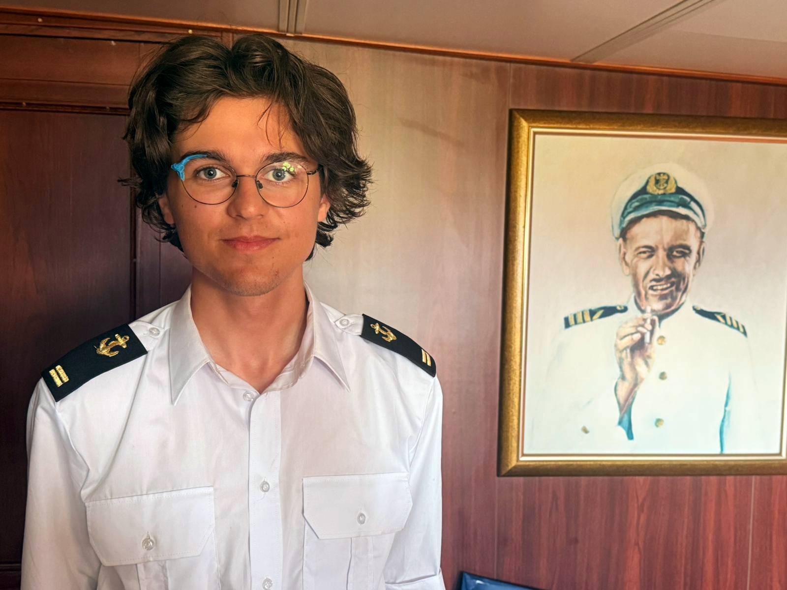 Portret młodego mężczyzny w białym mundurze marynarskim na tle drewnianego wnętrza, obok wiszącego na ścianie obrazu przedstawiającego kapitana. Młody człowiek to Maciej Słodki praktykant z Daru Młodzieży w czasie rejsu na 250-lecie Stanów Zjednoczonych