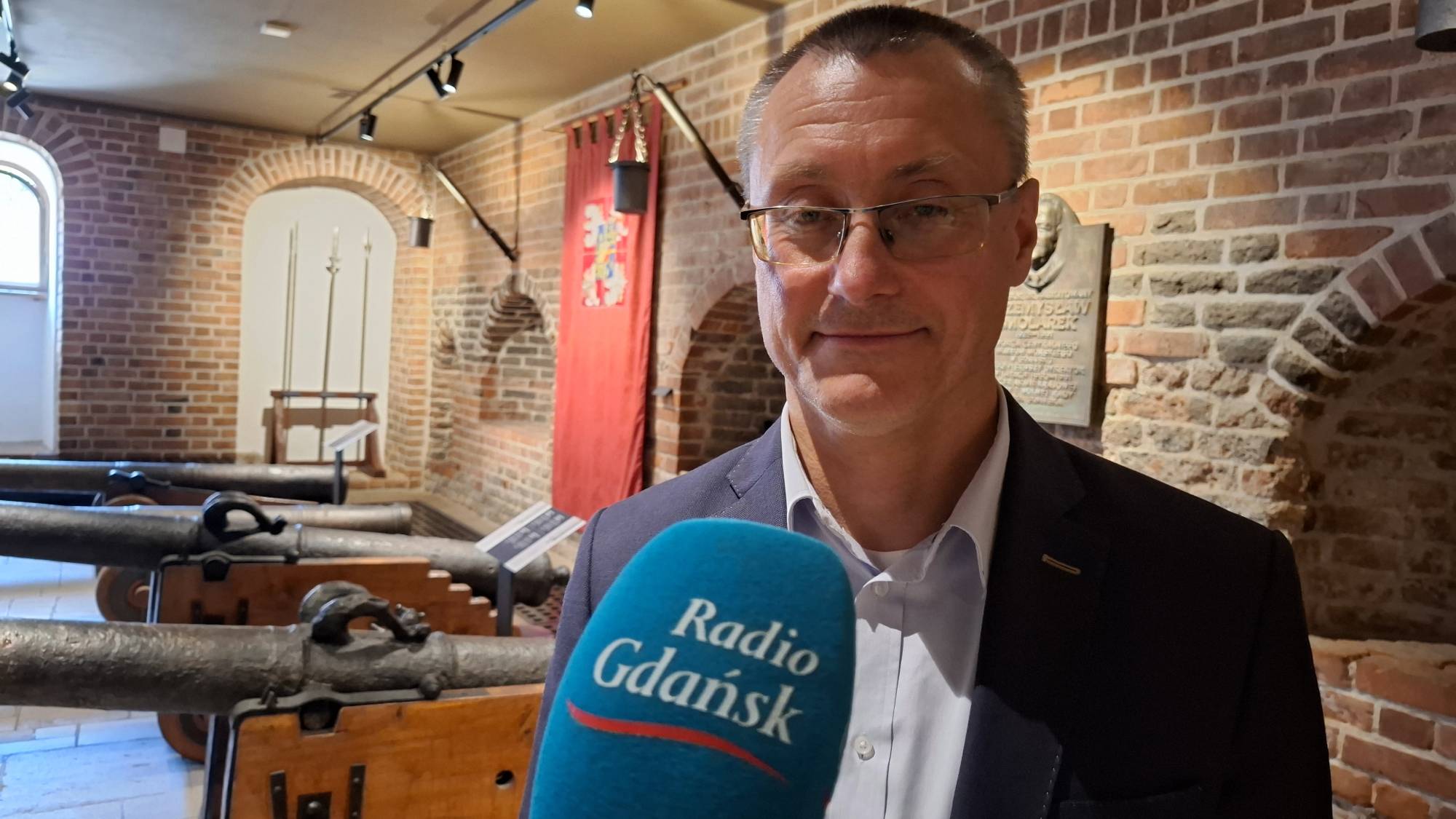 Mężczyzna w średnim wieku, w garniturze i okularach, z mikrofonem „Radio Gdańsk” przed sobą. Za nim widoczne są ściany z czerwonej cegły i kilka starych armat leżących na drewnianych podstawach. Postać na zdjęciu to Marcin Westphal, zastępca dyrektora Narodowego Muzeum Morskiego w Gdańsku