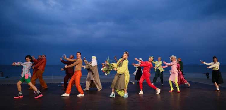 Aktorzy spektaklu „Na Waleta" w barwnych, widowiskowych kostiumach, pozujący w dynamicznych gestach na Scenie Letniej w Gdyni-Orłowie. W tle widoczne jest morze, plaża oraz ciemne, zachmurzone niebo o zmierzchu