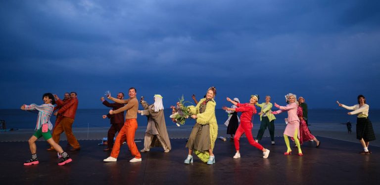Aktorzy spektaklu „Na Waleta" w barwnych, widowiskowych kostiumach, pozujący w dynamicznych gestach na Scenie Letniej w Gdyni-Orłowie. W tle widoczne jest morze, plaża oraz ciemne, zachmurzone niebo o zmierzchu