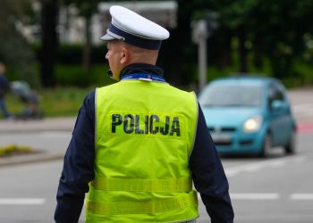 Umundurowany policjant w białej czapce z daszkiem i żółtej kamizelce z napisem "POLICJA" kieruje ruchem drogowym w centrum Gdańska. Stoi tyłem, zwrócony twarzą w lewo. W tle widać samochód osobowy wjeżdżający na skrzyżowanie dróg w Gdańsku. W mieście będą obowiązywać zmiany w organizacji w ruchu w związku z Konferencją na rzecz Odbudowy Ukrainy