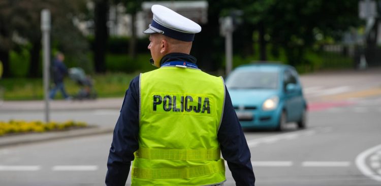 Umundurowany policjant w białej czapce z daszkiem i żółtej kamizelce z napisem "POLICJA" kieruje ruchem drogowym w centrum Gdańska. Stoi tyłem, zwrócony twarzą w lewo. W tle widać samochód osobowy wjeżdżający na skrzyżowanie dróg w Gdańsku. W mieście będą obowiązywać zmiany w organizacji w ruchu w związku z Konferencją na rzecz Odbudowy Ukrainy