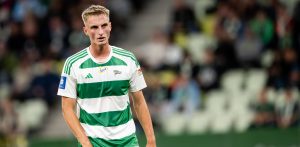 Elias Olsson, młody blondwłosy piłkarz w biało-zielonej, pasiastej koszulce Lechii Gdańsk, spoglądający w bok na tle rozmytych trybun stadionu