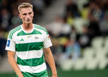 Elias Olsson, młody blondwłosy piłkarz w biało-zielonej, pasiastej koszulce Lechii Gdańsk, spoglądający w bok na tle rozmytych trybun stadionu