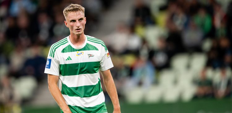 Elias Olsson, młody blondwłosy piłkarz w biało-zielonej, pasiastej koszulce Lechii Gdańsk, spoglądający w bok na tle rozmytych trybun stadionu