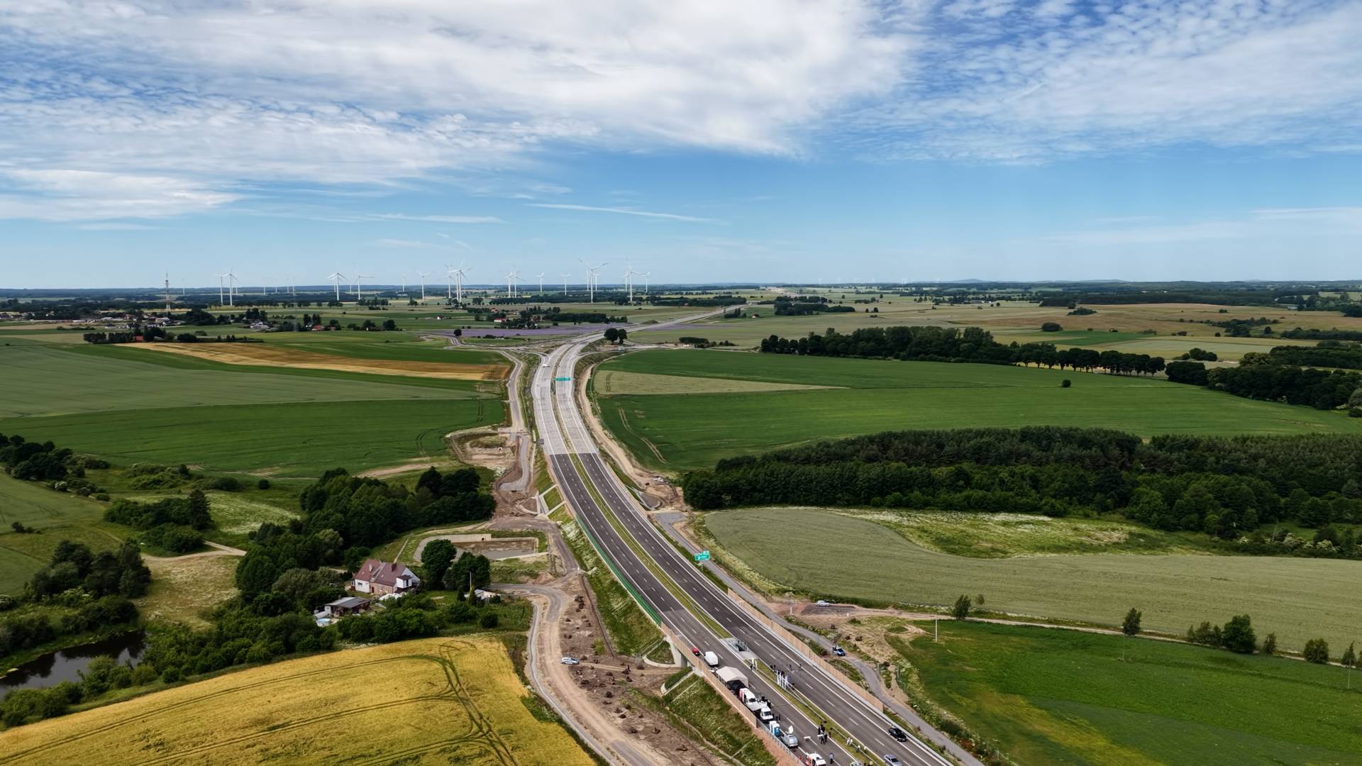 Wysoki widok z lotu ptaka na nowo wybudowaną drogę ekspresową S6 (Trasę Kaszubską) przecinającą rozległy, nizinny krajobraz. Dwujezdniowa trasa z ciemną nawierzchnią asfaltową biegnie środkiem kadru ku horyzontowi, lekko zakręcając w oddali. W dolnej części obrazu, na prawej jezdni, tuż przed przejściem nad drogą lokalną, widoczne jest zgrupowanie stojących pojazdów (głównie białych furgonów i aut osobowych).Po lewej stronie drogi, w dolnej części, znajduje się kilka domów jednorodzinnych, mały staw otoczony drzewami oraz pas żółtego pola uprawnego. Dalej rozciągają się rozległe, zielone pola, a na horyzoncie po lewej stronie widoczna jest farma wiatrowa z białymi turbinami. Po prawej stronie drogi dominują zielone uprawy polne przeplatane niewielkimi lasami i zagajnikami. Nad całym krajobrazem roztacza się jasnoniebieskie niebo pokryte gęstą siecią białych, pierzastych chmur