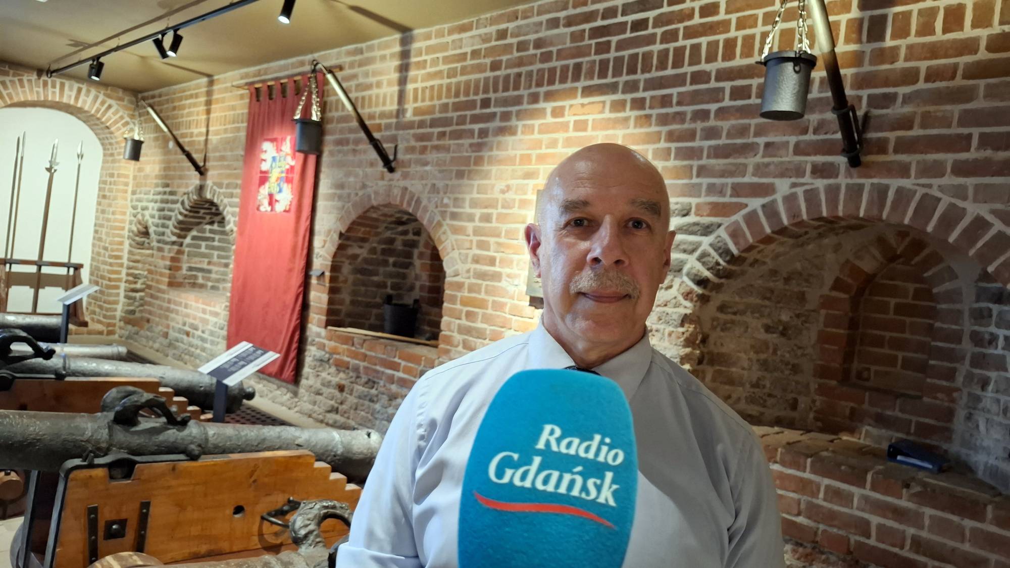 Łysy mężczyzna w średnim wieku, z wąsem, ubrany w jasną koszulę, przed sobą ma mikrofon z logo „Radio Gdańsk”. Za nim widoczne są ceglane ściany w starym budynku i kilka zabytkowych armat. Postać na zdjęciu to Piotr Stocki z Towarzystwa Przyjaciół Sołdka, statku-muzeum