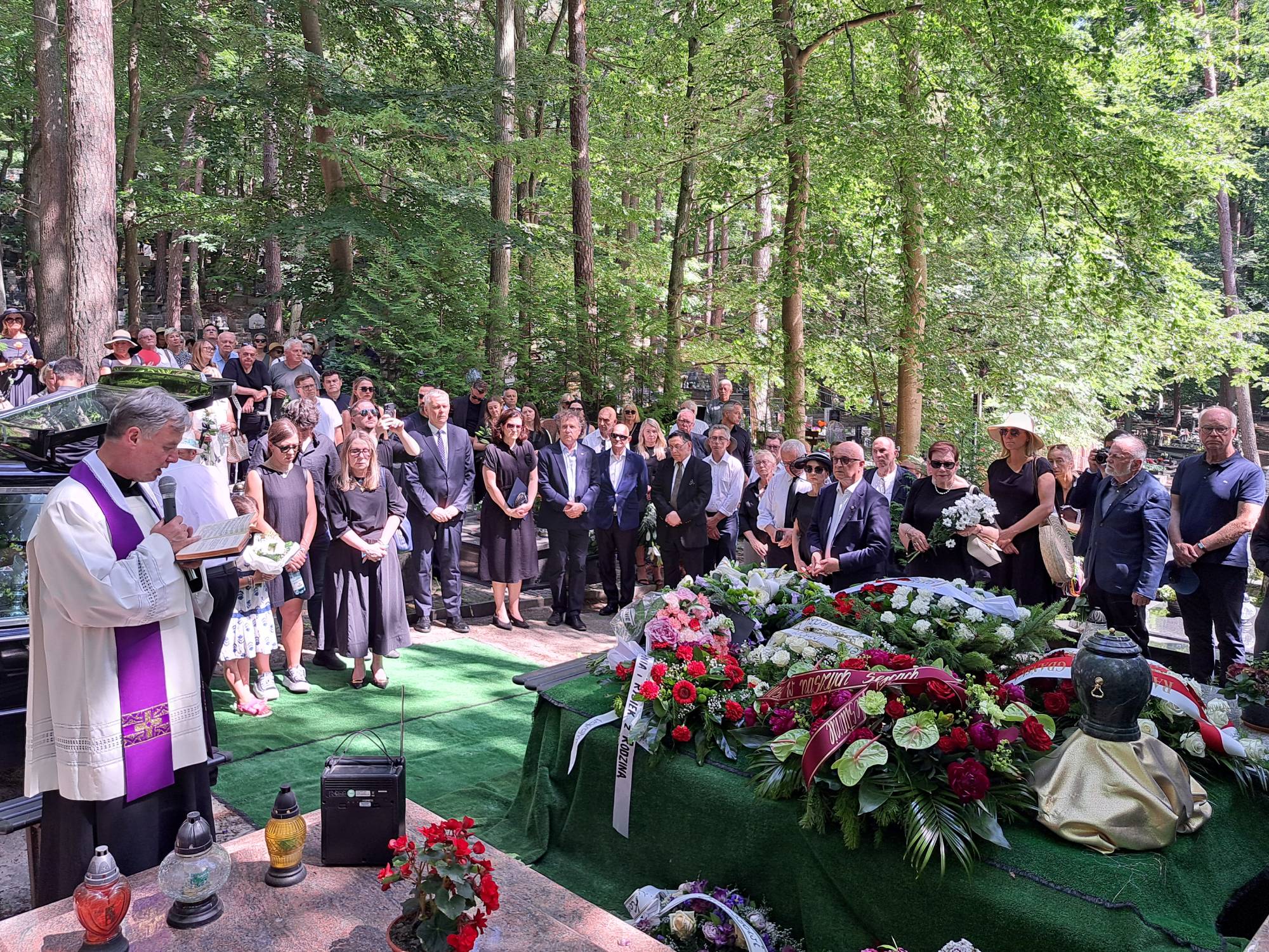 Pogrzeb Janusza Daszczyńskiego, byłego prezesa Telewizji Polskiej. Żałobnicy zgrupowani przy grobie, po lewej kapłan prowadzący uroczystość, w tle las