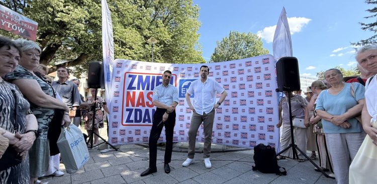 Patryk Jaki i Tomasz Bocheński stoją na placu w Słupsku przed dużym białym banerem z hasłem „Zmień nasze zdanie”. Politycy są otoczeni przez grupę mieszkańców obserwujących spotkanie.