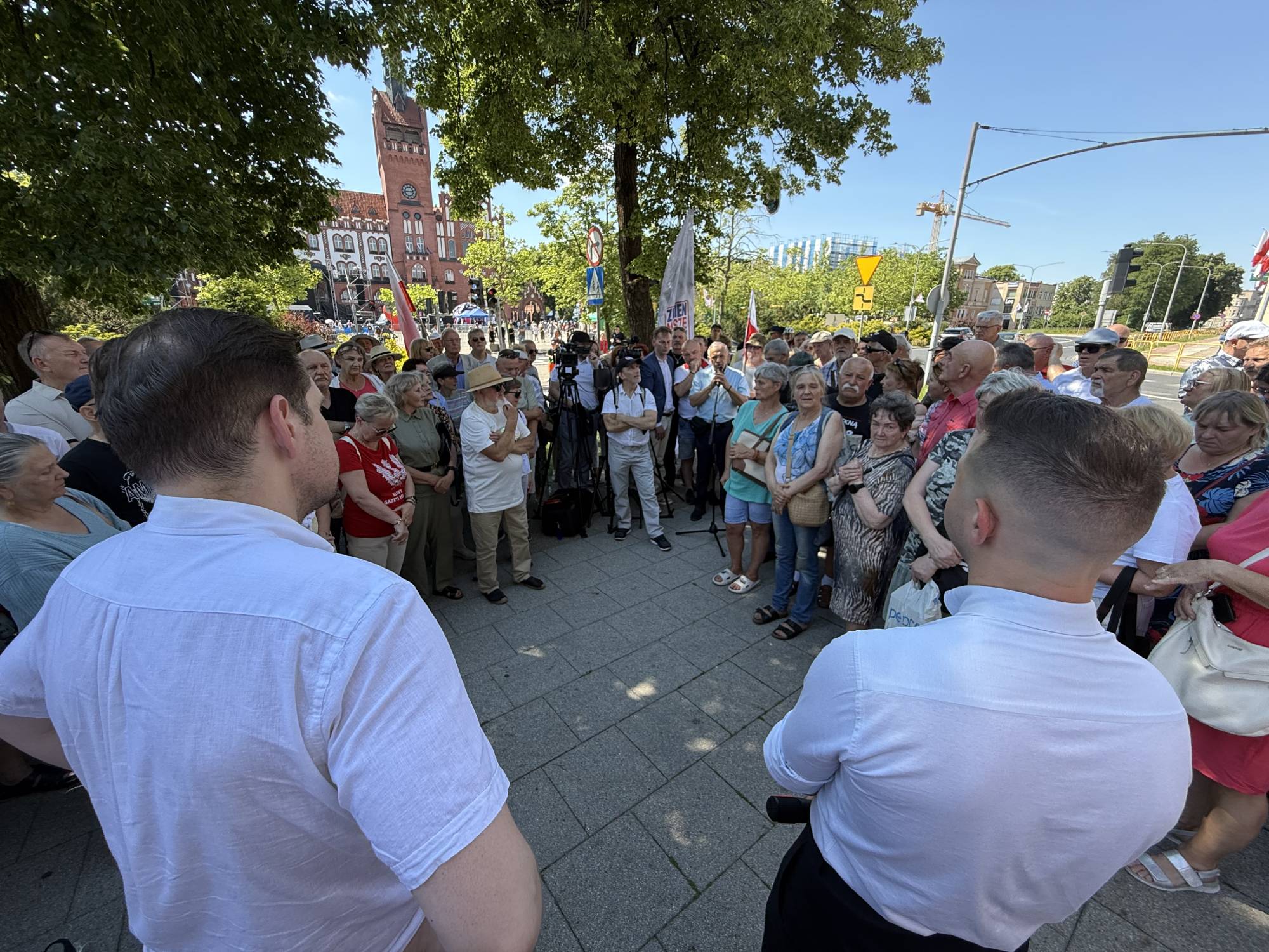 Zdjęcie wykonane z perspektywy zza pleców dwóch mężczyzn w białych koszulach (Tomasza Bocheńskiego po lewej i Patryka Jakiego po prawej), którzy stoją na wybrukowanym placu. Przed nimi, w półkolu, gromadzi się kilkadziesiąt osób słuchających wystąpienia. Spotkanie odbywa się w słoneczny dzień w otoczeniu drzew. W tle, za zgromadzonym tłumem, widać ulicę oraz charakterystyczny czerwony gmach ratusza w Słupsku z wysoką wieżą zegarową.