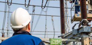 Polskie Sieci Elektroenergetyczne podpisały z umowę na rozbudowę stacji Gdańsk Błonia. Mężczyzna w białym kasku ochronnym i niebieskim stroju roboczym, widziany od tyłu, spogląda na infrastrukturę stacji elektroenergetycznej. Po prawej stronie, na podnośniku koszowym, inny pracownik wykonuje prace przy dużym, pionowym izolatorze wysokiego napięcia. W tle widoczne są stalowe konstrukcje, napowietrzne przewody oraz zielona tabliczka ostrzegawcza.