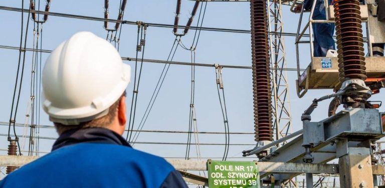 Polskie Sieci Elektroenergetyczne podpisały z umowę na rozbudowę stacji Gdańsk Błonia. Mężczyzna w białym kasku ochronnym i niebieskim stroju roboczym, widziany od tyłu, spogląda na infrastrukturę stacji elektroenergetycznej. Po prawej stronie, na podnośniku koszowym, inny pracownik wykonuje prace przy dużym, pionowym izolatorze wysokiego napięcia. W tle widoczne są stalowe konstrukcje, napowietrzne przewody oraz zielona tabliczka ostrzegawcza.