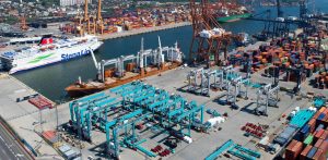 Widok z lotu ptaka na infrastrukturę terminalu w Porcie Gdynia. Po lewej stronie widoczny jest zacumowany prom pasażerski Stena Line oraz statek towarowy, w centrum stoją błękitne suwnice placowe, a po prawej rzędy kolorowych kontenerów i pomarańczowe suwnice nabrzeżowe. Ilustracja rozbudowy Portu Zewnętrznego w modelu Partnerstwa Publiczno-Prywatnego