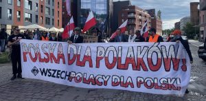 Protest przeciwko paktowi migracyjnemu w Gdańsku. Grupa osób protestuje na ulicy. Protestujący trzymają duży biały transparent z napisem "Polska dla Polaków" wykonanym dużymi czerwonymi literami. Poniżej mniejszymi czarnymi literami jest napisane "Wszechpolacy dla Polski"