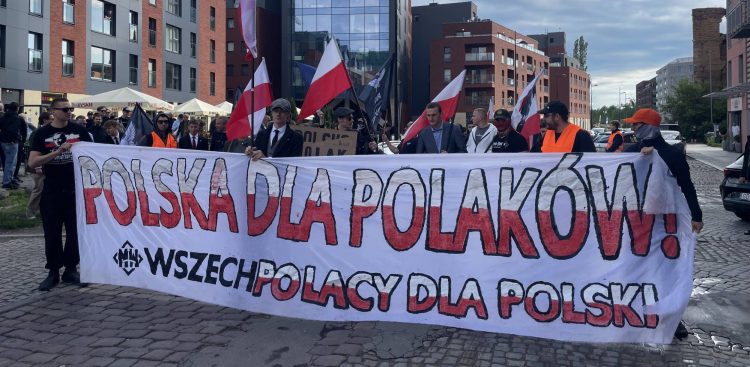 Protest przeciwko paktowi migracyjnemu w Gdańsku. Grupa osób protestuje na ulicy. Protestujący trzymają duży biały transparent z napisem "Polska dla Polaków" wykonanym dużymi czerwonymi literami. Poniżej mniejszymi czarnymi literami jest napisane "Wszechpolacy dla Polski"