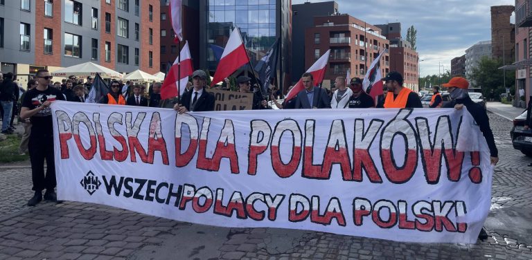 Protest przeciwko paktowi migracyjnemu w Gdańsku. Grupa osób protestuje na ulicy. Protestujący trzymają duży biały transparent z napisem "Polska dla Polaków" wykonanym dużymi czerwonymi literami. Poniżej mniejszymi czarnymi literami jest napisane "Wszechpolacy dla Polski"