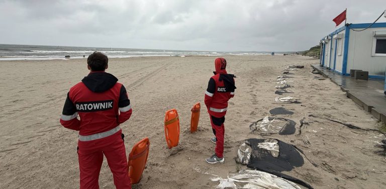 Ratownicy wodni na plaży wschodniej w Ustce. Dwóch ratowników w czerwono-czarnych strojach z napisem "RATOWNIK" na plecach. Stoją na piaszczystej plaży i patrzą w stronę morza. Obok nich znajdują się pomarańczowe boje ratunkowe. W oddali biało-niebieski budynek z czerwoną flagą na dachu. Plaża jest opustoszała, a w tle widać linię horyzontu z morzem, niebo jest zachmurzone. To początek sezonu kąpielowego