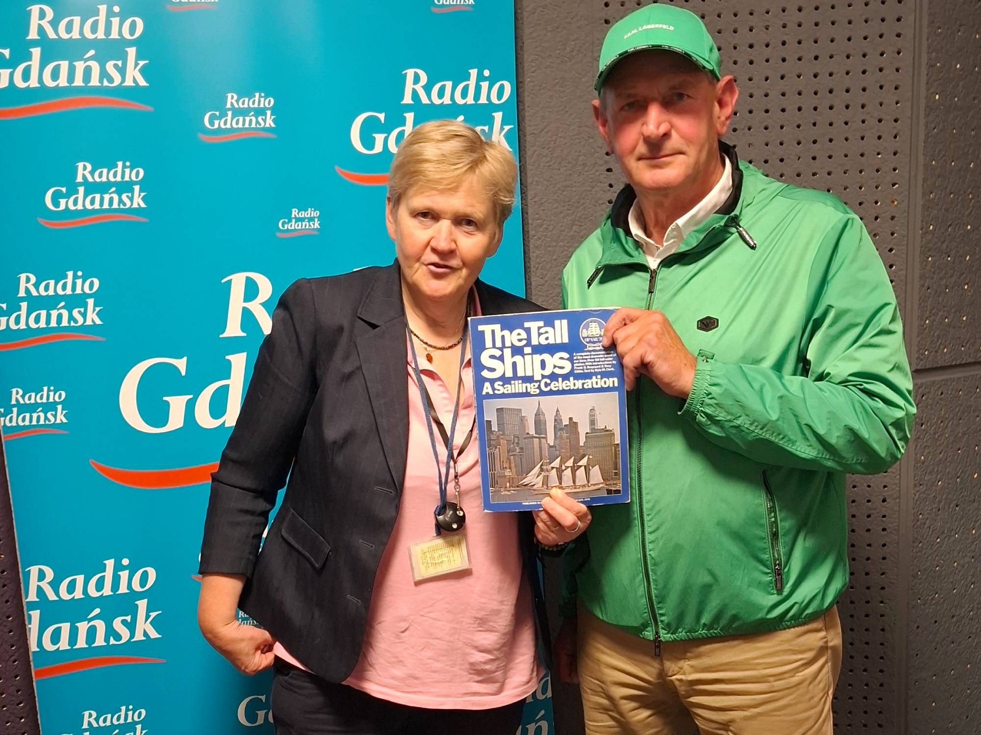 Kobieta w ciemnej marynarce i mężczyzna w zielonej kurtce oraz zielonej czapce z daszkiem stoją obok siebie na tle niebieskiej ścianki reklamowej z logo „Radio Gdańsk”. Wspólnie trzymają i pokazują do aparatu album zatytułowany „The Tall Ships: A Sailing Celebration”. To redaktor Anna Rębas z Radia Gdańsk oraz Kapitan Maciej Polarczyk, uczestnik historycznego rejsu Daru Pomorza na 200-lecie Stanów Zjednoczonych 