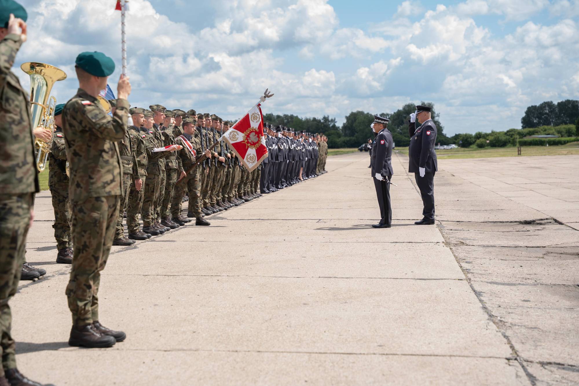 Święto 49. Bazy Lotniczej w Pruszczu Gdańskim. Uroczystość wojskowa na świeżym powietrzu. Na pierwszym planie, po lewej stronie, stoi grupa żołnierzy w mundurach polowych z instrumentami dętymi. Za nimi rozciąga się długi szereg żołnierzy w mundurach polowych trzymających karabiny. W centralnej części kadru widoczny jest poczet sztandarowy z dwoma sztandarami. Dalej, po prawej stronie, stoi dwóch oficerów w galowych mundurach, jeden z nich oddaje honor. W tle rozpościera się szeroka przestrzeń lotniska z trawą i lasem na horyzoncie, a nad nimi błękitne niebo z białymi chmurami