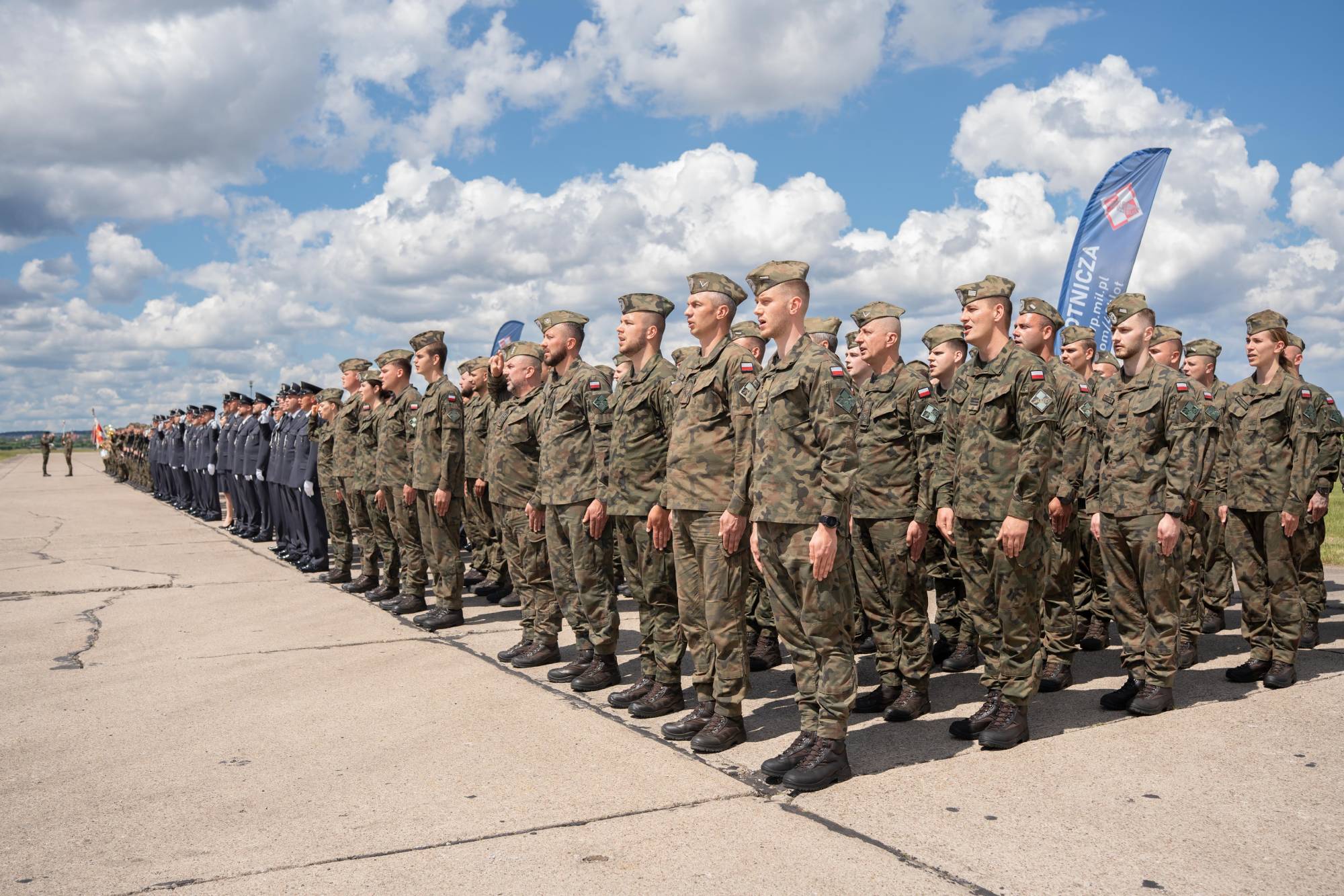 Święto 49. Bazy Lotniczej w Pruszczu Gdańskim. Żołnierze w mundurach polowych stoją w szyku na płycie lotniska podczas uroczystości wojskowej. W tle widoczny niebieski sztandar i błękitne niebo z chmurami