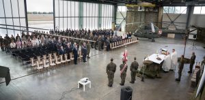 Święto 49. Bazy Lotniczej w Pruszczu Gdańskim. Uroczysty apel odbywający się w dużym hangarze lotniczym. W tle widoczny jest wojskowy śmigłowiec w barwach polskiego lotnictwa, częściowo zakryty siatką maskującą. Po lewej stronie zgromadzili się liczni żołnierze w mundurach, a także cywilni goście, siedzący na krzesłach. Po prawej stronie, przy ołtarzu nakrytym haftowanym obrusem stoi kilku kapłanów w białych szatach liturgicznych. Przed ołtarzem, tyłem do obiektywu, stoi kilku żołnierzy. Na pierwszym planie po lewej stronie widać stolik z projektorem. Przez otwartą bramę hangaru roztacza się widok na rozległe, zielone tereny