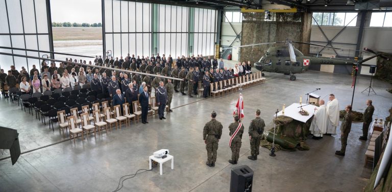 Święto 49. Bazy Lotniczej w Pruszczu Gdańskim. Uroczysty apel odbywający się w dużym hangarze lotniczym. W tle widoczny jest wojskowy śmigłowiec w barwach polskiego lotnictwa, częściowo zakryty siatką maskującą. Po lewej stronie zgromadzili się liczni żołnierze w mundurach, a także cywilni goście, siedzący na krzesłach. Po prawej stronie, przy ołtarzu nakrytym haftowanym obrusem stoi kilku kapłanów w białych szatach liturgicznych. Przed ołtarzem, tyłem do obiektywu, stoi kilku żołnierzy. Na pierwszym planie po lewej stronie widać stolik z projektorem. Przez otwartą bramę hangaru roztacza się widok na rozległe, zielone tereny