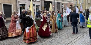 Uczestnicy święta ulicy Świętego Ducha w Gdańsku ubrani w stroje historyczne stoją na brukowanej ulicy. W tle stare kamienice