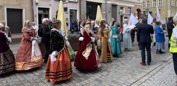 Uczestnicy święta ulicy Świętego Ducha w Gdańsku ubrani w stroje historyczne stoją na brukowanej ulicy. W tle stare kamienice