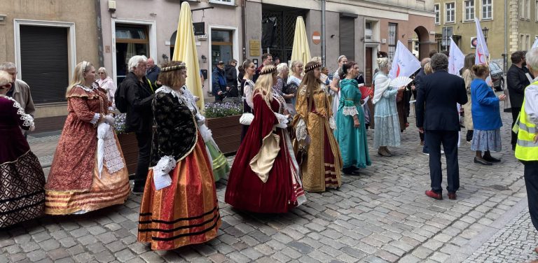 Uczestnicy święta ulicy Świętego Ducha w Gdańsku ubrani w stroje historyczne stoją na brukowanej ulicy. W tle stare kamienice