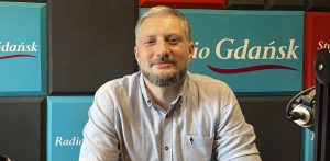 Uśmiechnięty mężczyzna w średnim wieku z krótkimi, lekko osiwiałymi włosami i zadbaną brodą. To Szymon Czajkowski, kierownik Zagrody Inicjatyw Twórczych w Swołowie. Ma na sobie jasnoszarą, zapinaną na guziki koszulę z długim rękawem. Siedzi w studiu nagraniowym - po prawej stronie kadru widoczny jest fragment profesjonalnego mikrofonu na ramieniu statywu. Tło stanowią kwadratowe panele akustyczne w kolorach turkusowym, czarnym i czerwonym, a na turkusowych widnieje biały napis „Radio Gdańsk”. Gość zapowiada festiwal EtnoBaltica