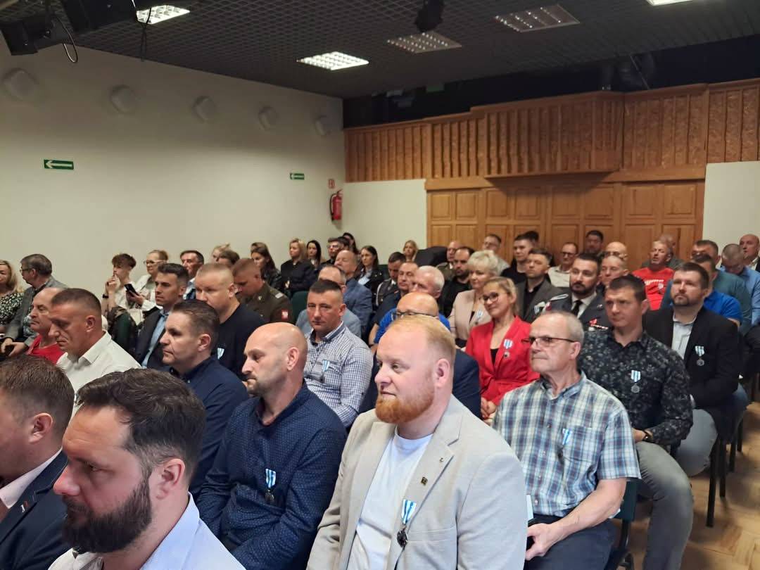 Uczestnicy uroczystości wręczenia medali „Honorowego Dawcy Krwi – Zasłużonego dla Zdrowia Narodu” w Słupsku siedzą na krzesłach w sali, słuchając przemówień. Wiele osób, zwłaszcza w pierwszych rzędach, ma przypięte medale do ubrań. W tle widać białe ściany oraz drewniane wykończenia