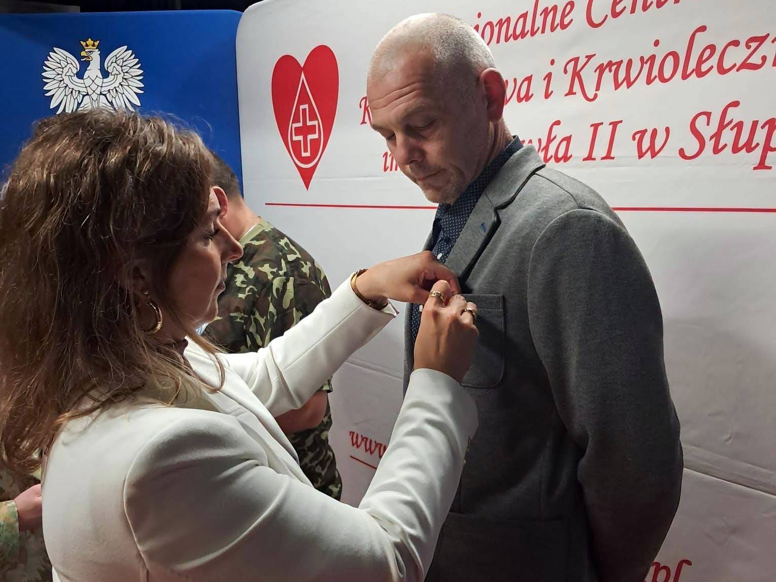 Uroczyste wręczenie medali „Honorowego Dawcy Krwi – Zasłużonego dla Zdrowia Narodu” w Słupsku. Kobieta w białej marynarce przypina medal do marynarki mężczyzny, który stoi przed nią, lekko pochylony. Oboje skupieni są na ceremonii. W tle widać fragmenty banerów z napisami, w tym logo z sercem i krzyżem.