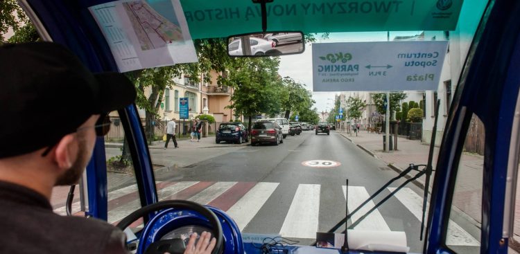 Widok z wnętrza niebieskiego eko-busa jadącego ulicą w stronę centrum Sopotu, przedstawiony z perspektywy pasażera siedzącego za kierowcą. Po lewej stronie widoczny jest profil kierowcy w czarnej czapce z daszkiem, trzymającego dłoń na niebieskiej kierownicy. Przez przednią szybę pojazdu widać asfaltową drogę z przejściem dla pieszych (zebrą) oraz namalowanym na jezdni ograniczeniem prędkości do 30. Na szybie naklejone są odwrócone tyłem (w odbiciu lustrzanym) tabliczki informacyjne, w tym jedna wskazująca drogę na bezpłatny eko-parking przy Ergo Arenie oraz do centrum i na plażę. W tle, wzdłuż drogi, ciągną się drzewa, chodnik z pieszymi oraz zaparkowane samochody na tle miejskich budynków