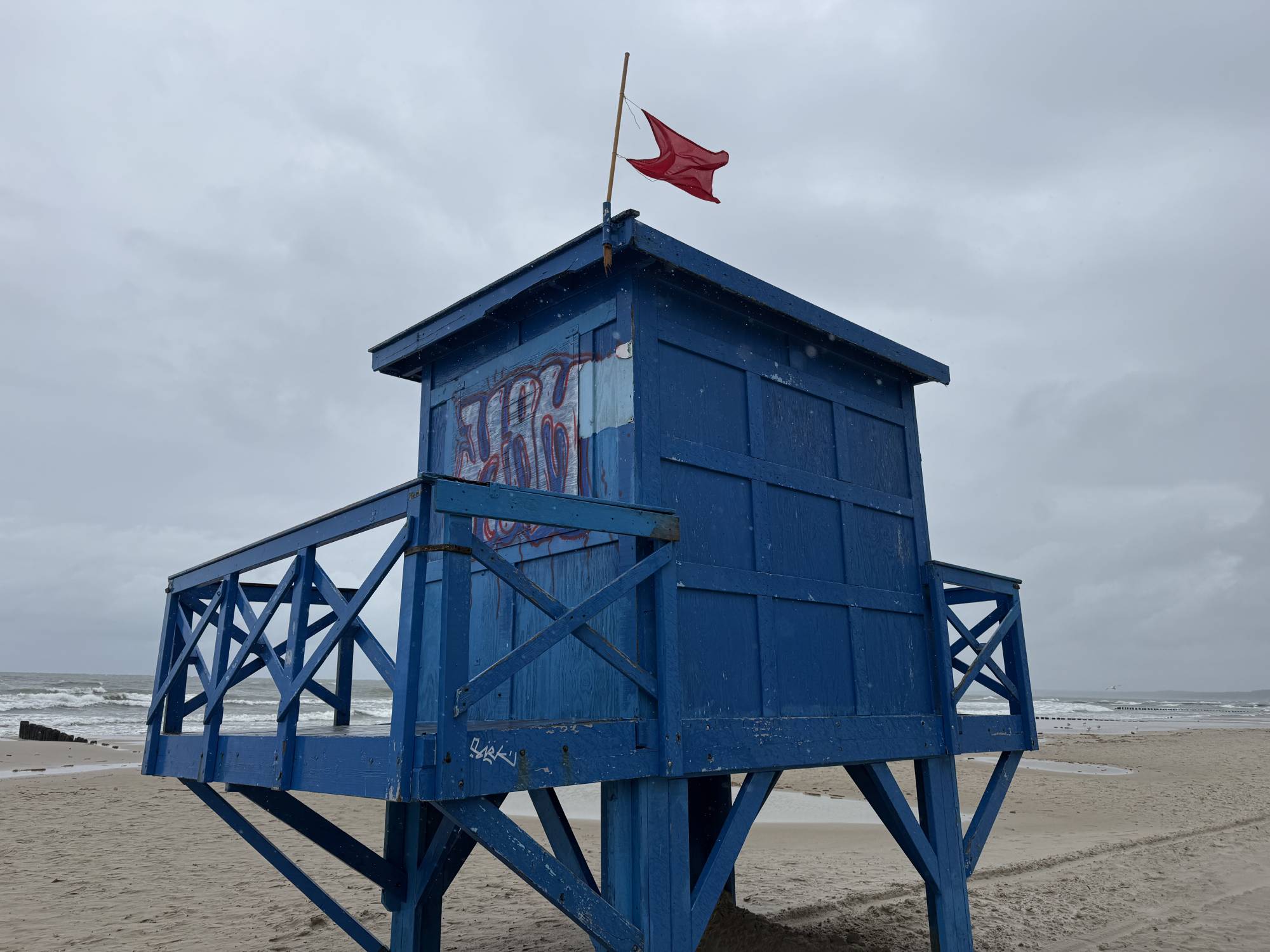 Niebieska wieża ratownicza stoi na piaszczystej plaży w Ustce pod pochmurnym niebem. Na dachu wieży powiewa czerwona flaga. Na ścianach wieży widać ślady graffiti. W tle rozciąga się morze, z widocznymi falami i fragmentem falochronu w oddali. Plaża jest pusta. To początek sezony kąpielowego