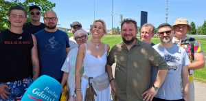 Grupa uśmiechniętych wolontariuszy fundacji „Zupa na Monciaku” pozuje do wspólnego zdjęcia w słoneczny, letni dzień. W kadrze znajduje się około dziesięciu osób – kobiet i mężczyzn w różnym wieku, ubranych w luźne, codzienne stroje. W lewym dolnym rogu widoczny jest niebieski mikrofon z logo „Radio Gdańsk”. W tle widać zielony teren trawiasty, barierki ochronne oraz elementy infrastruktury drogowej pod bezchmurnym, błękitnym niebem, co sugeruje przygotowania do podróży na świeżym powietrzu