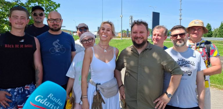 Grupa uśmiechniętych wolontariuszy fundacji „Zupa na Monciaku” pozuje do wspólnego zdjęcia w słoneczny, letni dzień. W kadrze znajduje się około dziesięciu osób – kobiet i mężczyzn w różnym wieku, ubranych w luźne, codzienne stroje. W lewym dolnym rogu widoczny jest niebieski mikrofon z logo „Radio Gdańsk”. W tle widać zielony teren trawiasty, barierki ochronne oraz elementy infrastruktury drogowej pod bezchmurnym, błękitnym niebem, co sugeruje przygotowania do podróży na świeżym powietrzu