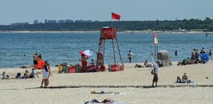 Słoneczny dzień na piaszczystej plaży nad morzem. W centralnej części kadru stoi czerwona, metalowa wieżyczka ratownicza, na której szczycie powiewa czerwona flaga oznaczająca zakaz kąpieli (tym razem z powodu sinic) Na plaży widoczni są wypoczywający na kocach ludzie oraz pojedyncze osoby spacerujące po drewnianej ścieżce wzdłuż piasku. W tle rozciąga się spokojna woda Zatoki Gdańskiej, a na horyzoncie widać zieloną linię drzew oraz zarysy miejskiej zabudowy