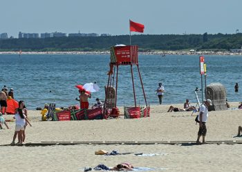 Słoneczny dzień na piaszczystej plaży nad morzem. W centralnej części kadru stoi czerwona, metalowa wieżyczka ratownicza, na której szczycie powiewa czerwona flaga oznaczająca zakaz kąpieli (tym razem z powodu sinic) Na plaży widoczni są wypoczywający na kocach ludzie oraz pojedyncze osoby spacerujące po drewnianej ścieżce wzdłuż piasku. W tle rozciąga się spokojna woda Zatoki Gdańskiej, a na horyzoncie widać zieloną linię drzew oraz zarysy miejskiej zabudowy